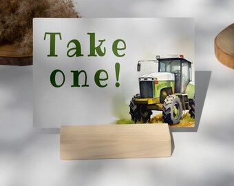 Puede incluir: Una tarjeta blanca con texto verde que dice "¡Toma uno!" y una ilustración de acuarela de un tractor verde y blanco. La tarjeta está apoyada en un soporte de madera.
