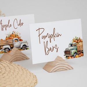 May include: Two white cards with brown text and illustrations of a vintage truck filled with pumpkins. The text on the cards reads "Apple Cider" and "Pumpkin Bars". The cards are propped up on small wooden stands.