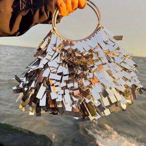 May include: A silver handbag with a circular handle. The bag is covered in rectangular, reflective pieces that create a shimmering effect. The bag is held up against a backdrop of the ocean and sky.