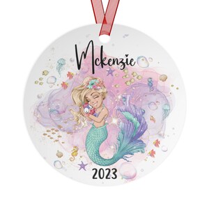 May include: A round, white ornament with a pink and purple watercolor background. A mermaid with blonde hair and a teal tail is holding a small, pink fish. The ornament has gold glitter and the text "Mckenzie 2023".