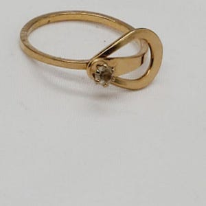 May include: A gold ring with a single clear stone set in the center of the band. The band is a simple, thin band with a unique design that wraps around the finger.