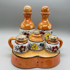 May include: A ceramic condiment set featuring two oil or vinegar bottles with stoppers, two salt shakers, and a sugar bowl with a spoon. The set is decorated with floral patterns and sits on a matching tray.