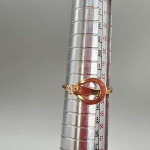 May include: A gold ring with a clear stone set in a double-loop design. The ring is sized on a metal ring sizer, showing a size of 52.