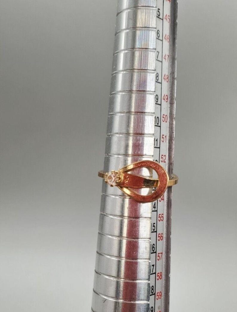 May include: A gold ring with a clear stone set in a double-loop design. The ring is sized on a metal ring sizer, showing a size of 52.