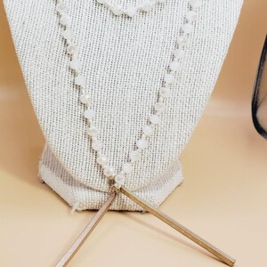 Puede incluir: Un collar de varias capas con cuentas blancas facetadas y un colgante de barra dorada. El collar se exhibe sobre un busto de lino blanco sobre un fondo neutro. Las barras del colgante miden aproximadamente 7.6 cm.