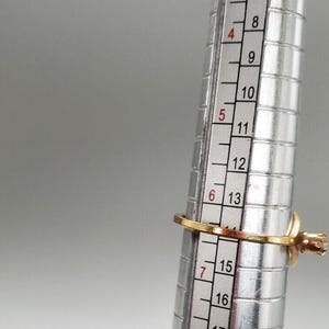 May include: A gold ring with a single clear stone is being sized on a metal ring sizer. The ring size is 7.5.