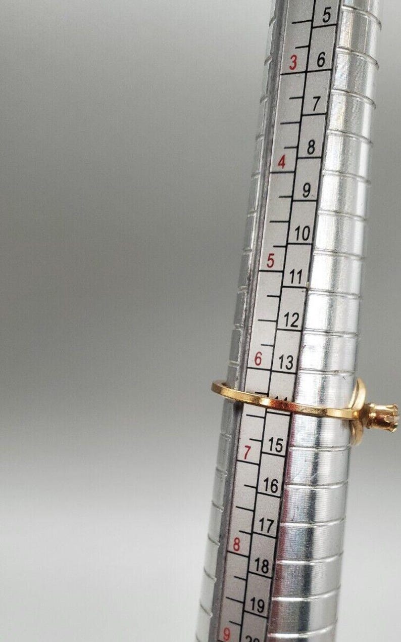May include: A gold ring with a single clear stone is being sized on a metal ring sizer. The ring size is 7.5.