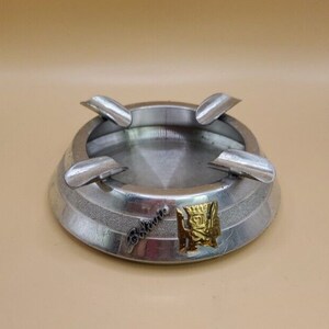 May include: A silver ashtray with four slots for cigarettes. The ashtray has a gold emblem with a crown and a shield on it. The text "Palatino" is engraved on the ashtray.
