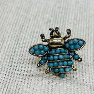 May include: A gold bee brooch with turquoise blue beads. The bee has a detailed design with black eyes and raised wings.