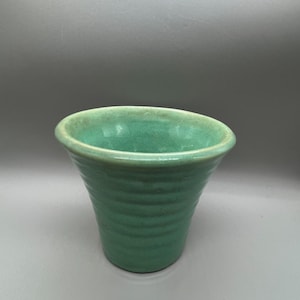 Op de afbeelding: Een kleine, uitlopende keramische beker met een levendig turquoise glazuur. De buitenkant is voorzien van een reeks horizontale, geribbelde groeven die rond het lichaam lopen, terwijl de binnenkant en rand een gladde, lichtere blauwgroene tint hebben.