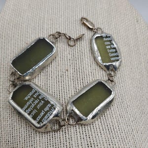 May include: Silver bracelet with four rectangular green glass charms. Each charm has text printed on it. The text on one charm reads: "S./Probado e sulle person t your loca de recyclag bus informe proche." The text on another charm reads: "To Avoid cou. F se Us Evitar Gesich verme"