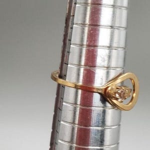 May include: A gold ring with a clear stone set in a heart-shaped design. The ring is on a metal ring sizer.
