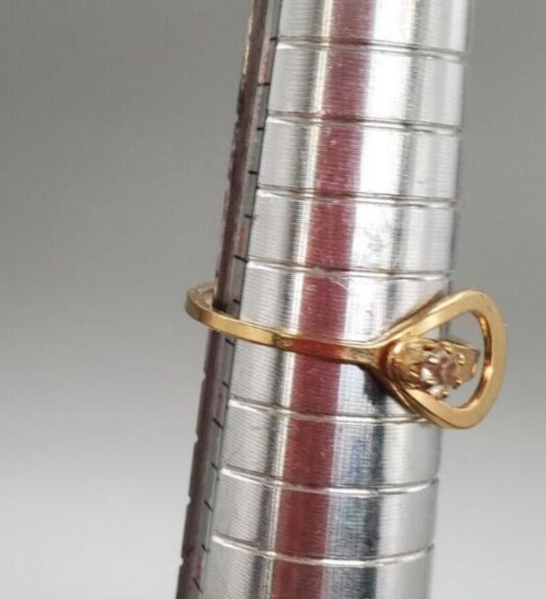 May include: A gold ring with a clear stone set in a heart-shaped design. The ring is on a metal ring sizer.
