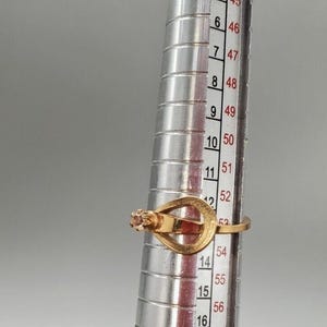 May include: A gold ring with a single pink stone set in the center. The ring is sized using a ring sizer, which shows the ring size is 54.