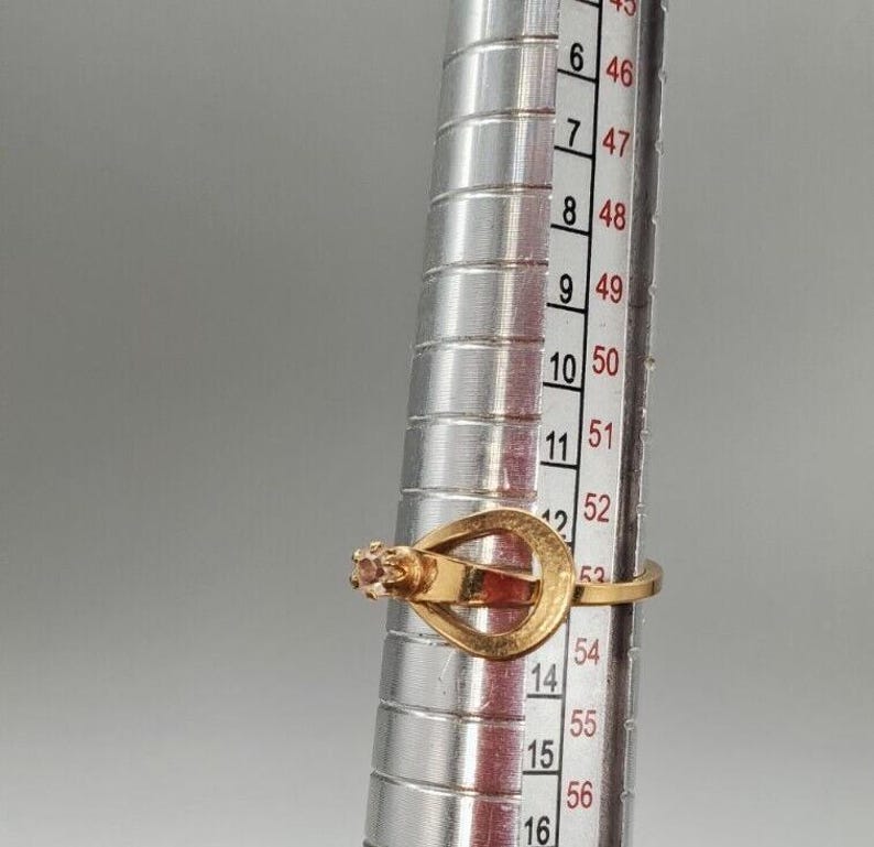 May include: A gold ring with a single pink stone set in the center. The ring is sized using a ring sizer, which shows the ring size is 54.