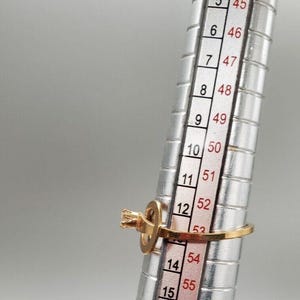 May include: A gold ring with a single clear stone set in a prong setting. The ring is sized on a metal ring sizer, showing a size of 14.