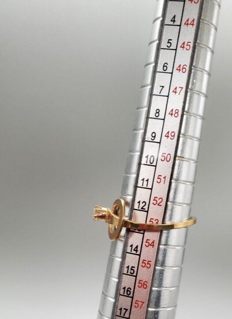May include: A gold ring with a single clear stone set in a prong setting. The ring is sized on a metal ring sizer, showing a size of 14.