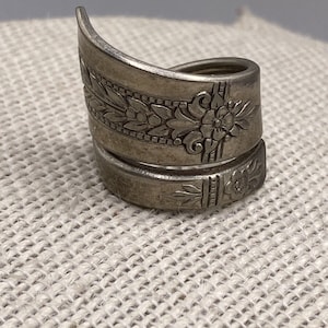Puede incluir: Un anillo de plata en forma de cuchara con un diseño floral. El anillo está hecho de una cuchara doblada y tiene un aspecto único y rústico.