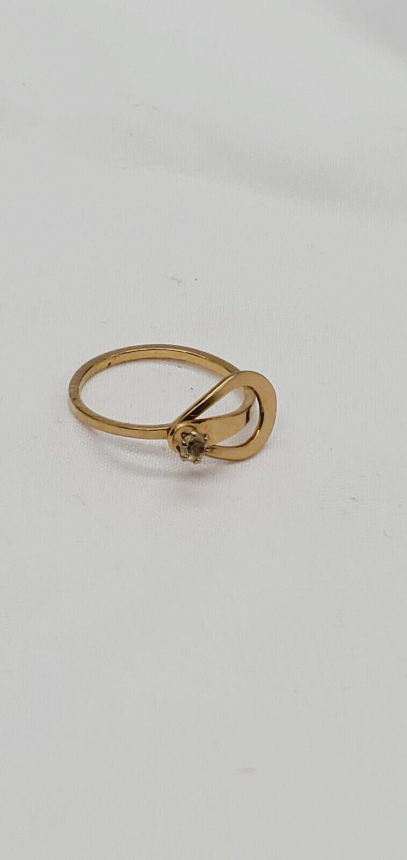 May include: A gold ring with a single small diamond set in the center. The ring has a unique design with two bands that intersect and form a loop.
