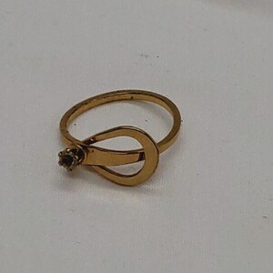 May include: A gold ring with a unique design featuring a small, clear stone. The ring is open and has a curved, teardrop-shaped design.