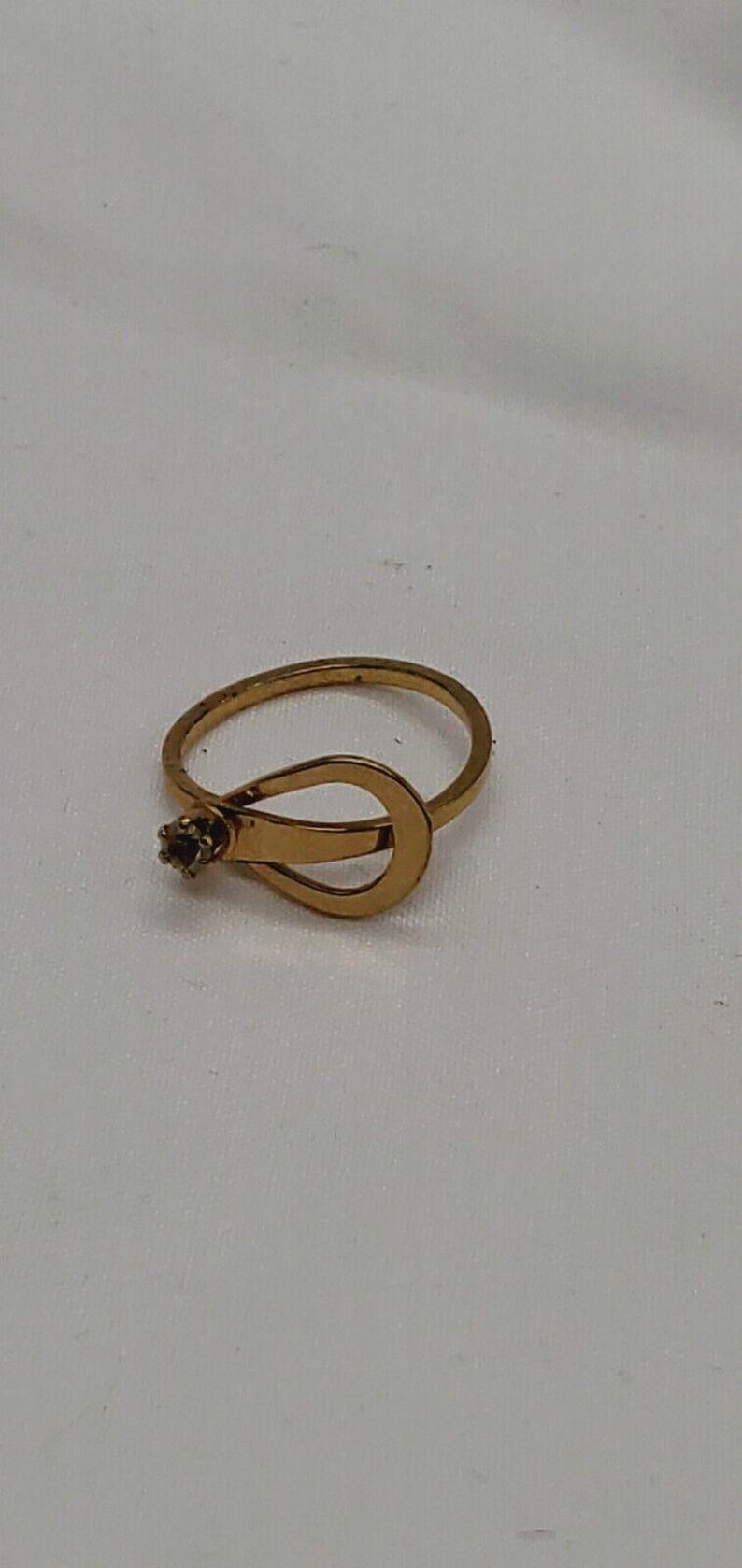 May include: A gold ring with a unique design featuring a small, clear stone. The ring is open and has a curved, teardrop-shaped design.
