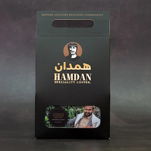 May include: Black Hamdan Specialty Coffee box with a handle, gold lettering, and an image of a man. The box has the text "Sipping Culture Building Community" at the top. A smaller window shows a picture of the coffee and a man.