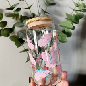 May include: A clear glass reusable water bottle with a bamboo lid and a pink butterfly design. The bottle has a glass straw.