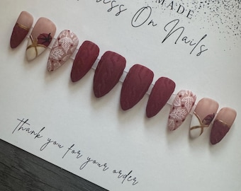 May include: Set of ten burgundy press-on nails with white floral designs and gold accents. The nails are arranged on a white card with the text "Handmade Press On Nails" and "Thank you for your order".