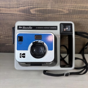 Puede incluir: Una cámara instantánea Kodak vintage, modelo "the Handle", en blanco y negro con un acento azul. La cámara tiene una lente negra, un visor y una correa negra. El texto "the Handle" y "A Kodak Instant Camera" son visibles.