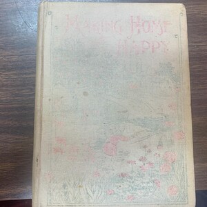 Puede incluir: Un libro vintage titulado "Making Home Happy" con un diseño floral en la portada. La portada es de color crema con ilustraciones florales rosas y verdes.