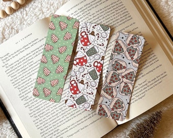 May include: Three colorful bookmarks with different designs. The first bookmark has a green and red pattern with a Christmas tree design. The second bookmark has a white background with a hot chocolate and candy cane design. The third bookmark has a gray background with a pizza design.