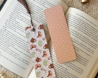 May include: A close-up of two bookmarks. One bookmark is pink with white polka dots. The other bookmark is a light brown with a pattern of teacups and teapots. The text on the open book reads "The Invisible Life of Addie LaRue".