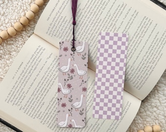 May include: Two decorative bookmarks rest on an open book. One bookmark features a lavender and white checkered pattern. The other has a lavender background with white geese and floral designs. A beaded strand lies nearby.