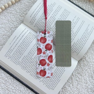 May include: A decorative bookmark featuring a red apple pattern with pink flowers and a red tassel. The bookmark is placed in an open book, with a second, green-checkered bookmark visible. The book is on a white, textured surface.