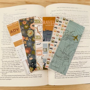 May include: A close-up of an open book with several colorful bookmarks. The bookmarks feature various designs, including floral patterns, travel-themed illustrations, and a map. The book's pages are visible, with text about a character named Henry.
