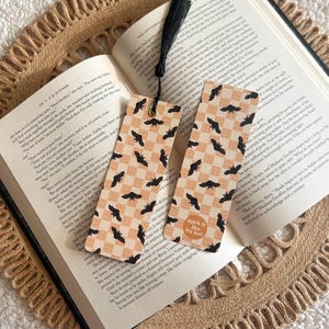 May include: Two rectangular bookmarks with a black bat pattern on a tan and white checkered background. Each bookmark has a black tassel. One bookmark has a sticker that says "mark it like it's hot".