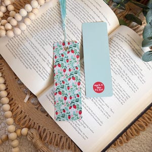 May include: Two bookmarks rest on an open book. One bookmark features a strawberry pattern with a teal tassel. The other is solid light blue with the text "mark it like it's hot" in a red circle. A wooden bead garland is in the background.
