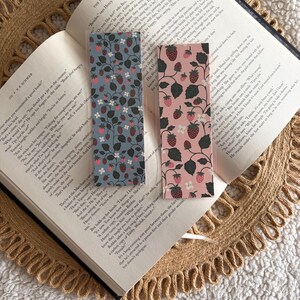 May include: Two rectangular bookmarks with a strawberry pattern rest on an open book. One bookmark is light blue, the other is pink. The book is open to a page with text.