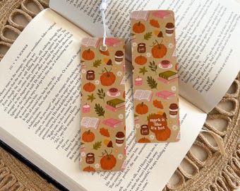 May include: Two decorative bookmarks with a fall theme, featuring pumpkins, coffee cups, books, and leaves on a tan background. One bookmark has the text "mark it like it's hot". A white tassel hangs from the top of one bookmark. The bookmarks are placed on an open book.