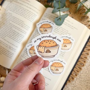 May include: A hand holding several stickers with a sourdough bread bowl design. The stickers feature a cartoon illustration of a bread bowl with a towel and the text "in my sourdough era". The stickers are on top of an open book.
