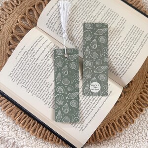 May include: Two decorative bookmarks rest on an open book. The bookmarks feature a repeating pattern of white seashells and starfish on a muted green background. One bookmark has a white tassel. The text "mark it like it's hot" is on one bookmark.