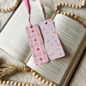 May include: Two pink and white patterned bookmarks with pink tassels. The bookmarks feature the text "Hot Girls Read" in a pink and white font. One bookmark has a pink and white checkered pattern with a pink flower. The other bookmark has a pink and white pattern with glasses, a cup of coffee, a book, and a candle.