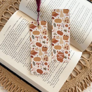 May include: Two rectangular bookmarks with a fall-themed pattern of pumpkins, leaves, and teapots. One bookmark has the text "mark it like it's hot." A burgundy tassel hangs from the top. The bookmarks are placed on an open book.