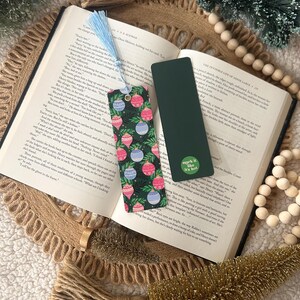 May include: Two bookmarks rest on an open book. One features a dark green background with colorful Christmas ornament designs and a blue tassel. The other is solid dark green with the text "mark it like it's hot."
