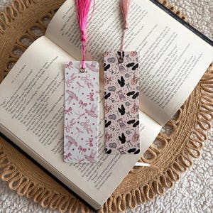 May include: Two decorative bookmarks with pink tassels rest on an open book. One bookmark features a pattern of ballet shoes, bows, and ribbons. The other has a pattern of ballet shoes and silhouettes. The book is on a woven placemat.