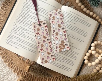 May include: Two festive bookmarks with a beige background and a repeating pattern of gingerbread men, sweaters, bows, and other holiday-themed icons. One bookmark has a burgundy tassel. The text "mark it like it's hot" is on one bookmark.