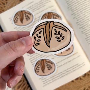 May include: A hand holding a sticker with a cartoon illustration of a loaf of bread. The sticker is light brown with darker brown details and leaf-like designs. Several similar stickers are scattered on an open book.