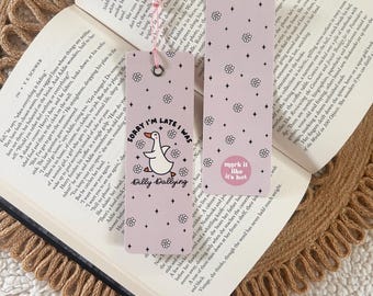 May include: Two pink bookmarks with tassel ends rest on an open book. One bookmark reads "Sorry I'm late I was Dilly Dallying" with a cartoon duck. The other says "mark it like it's hot" with floral accents.