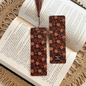 May include: Two rectangular bookmarks with a pumpkin and floral pattern on a dark brown background. One bookmark has the text "mark it like it's hot". A brown tassel is attached to the top. The bookmarks are placed on an open book.