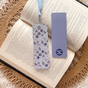 May include: A book is open to a page with text, with two bookmarks. One is a light blue rectangle with the text "mark it like it's hot". The other bookmark has a blue tassel and a pattern of seashells and starfish.